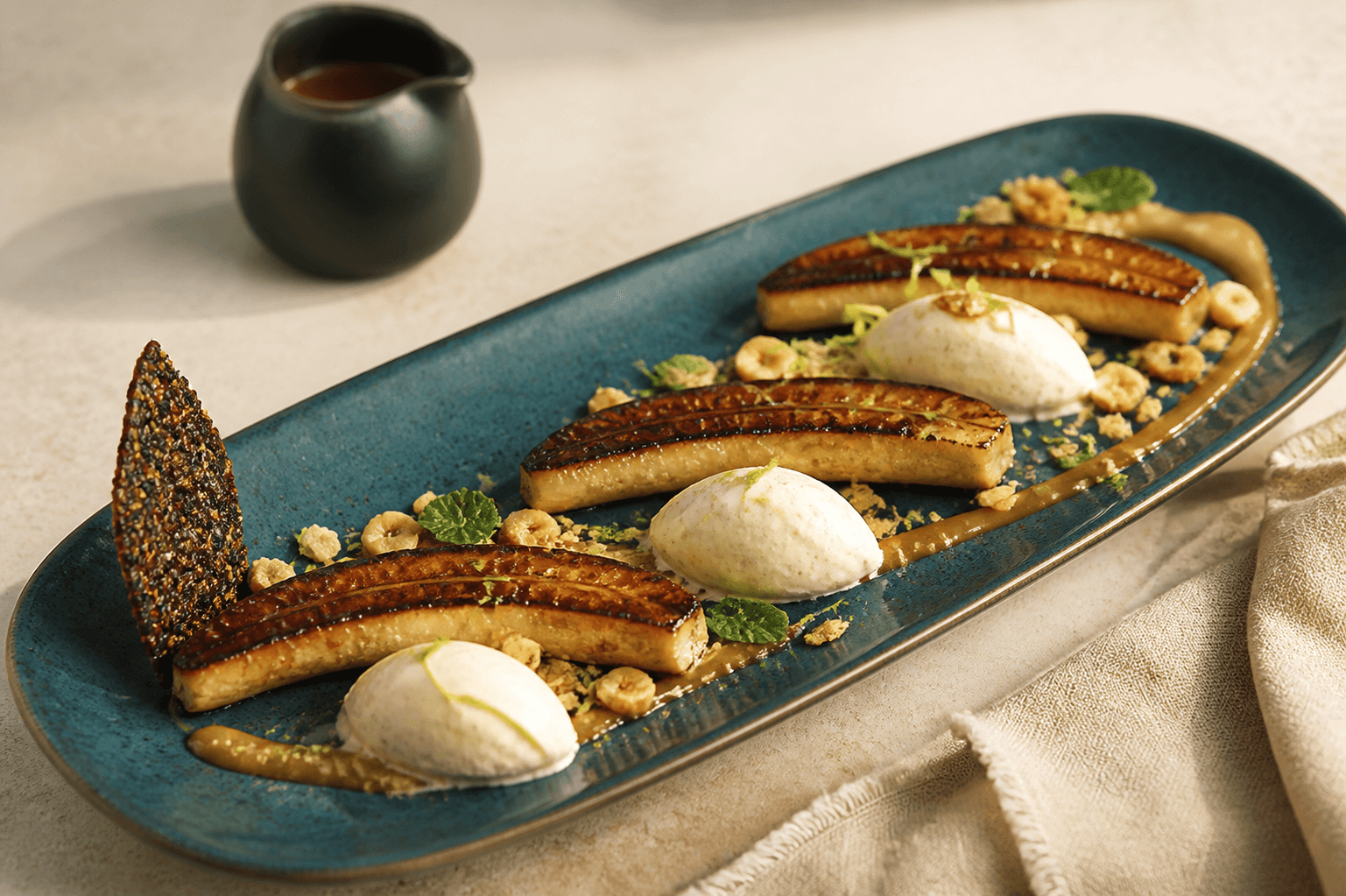 Tre grillede bananhalvdele med mørk rom-brunet smørkaramel, kærnemælks-cardamomis, hasselnøddeknas og kakaotuile på dybt petrolfad