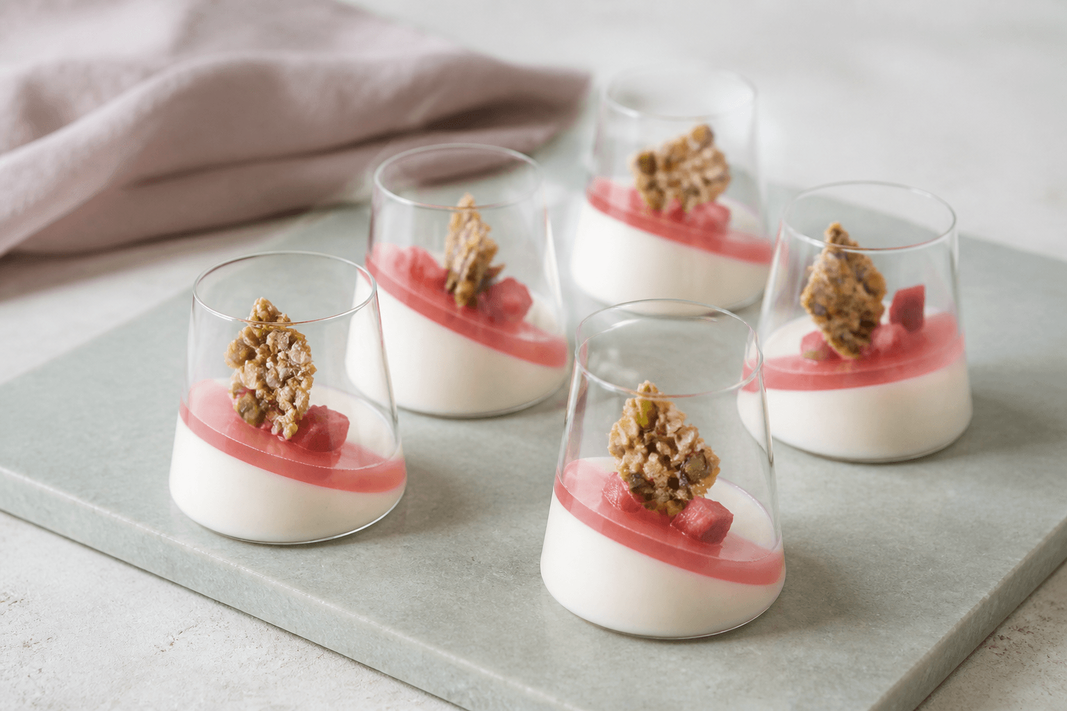 Fem rabarber-panna-cotta med kærnemælk, skråt pink gelélag, rabarbertern og pistacie-havreknas i lave koniske glas
