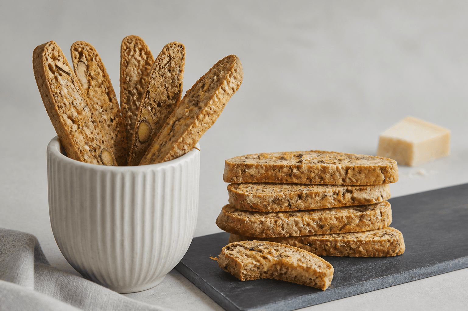 Lange parmesanbiscotti med rug, pistacie, citron, rosmarin og sort peber stående i hvid kop og stablet på sort skifer
