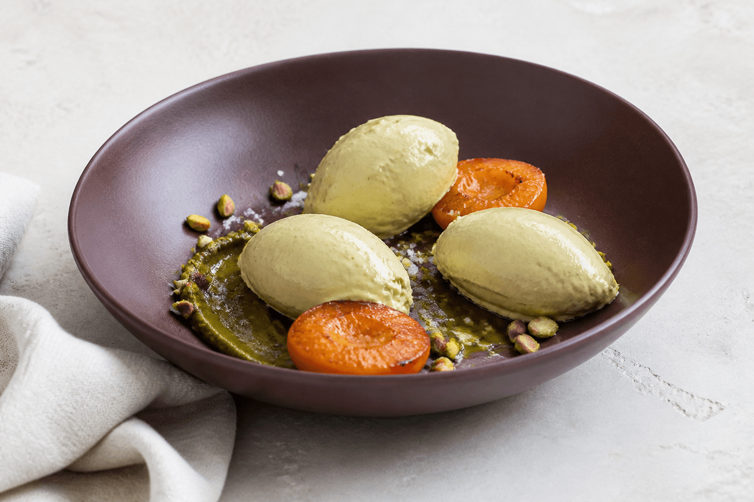 Tre queneller naturlig pistaciegelato med salt pistaciepraliné, ristede abrikoser, pistacier, olivenolie og flagesalt i bordeaux skål