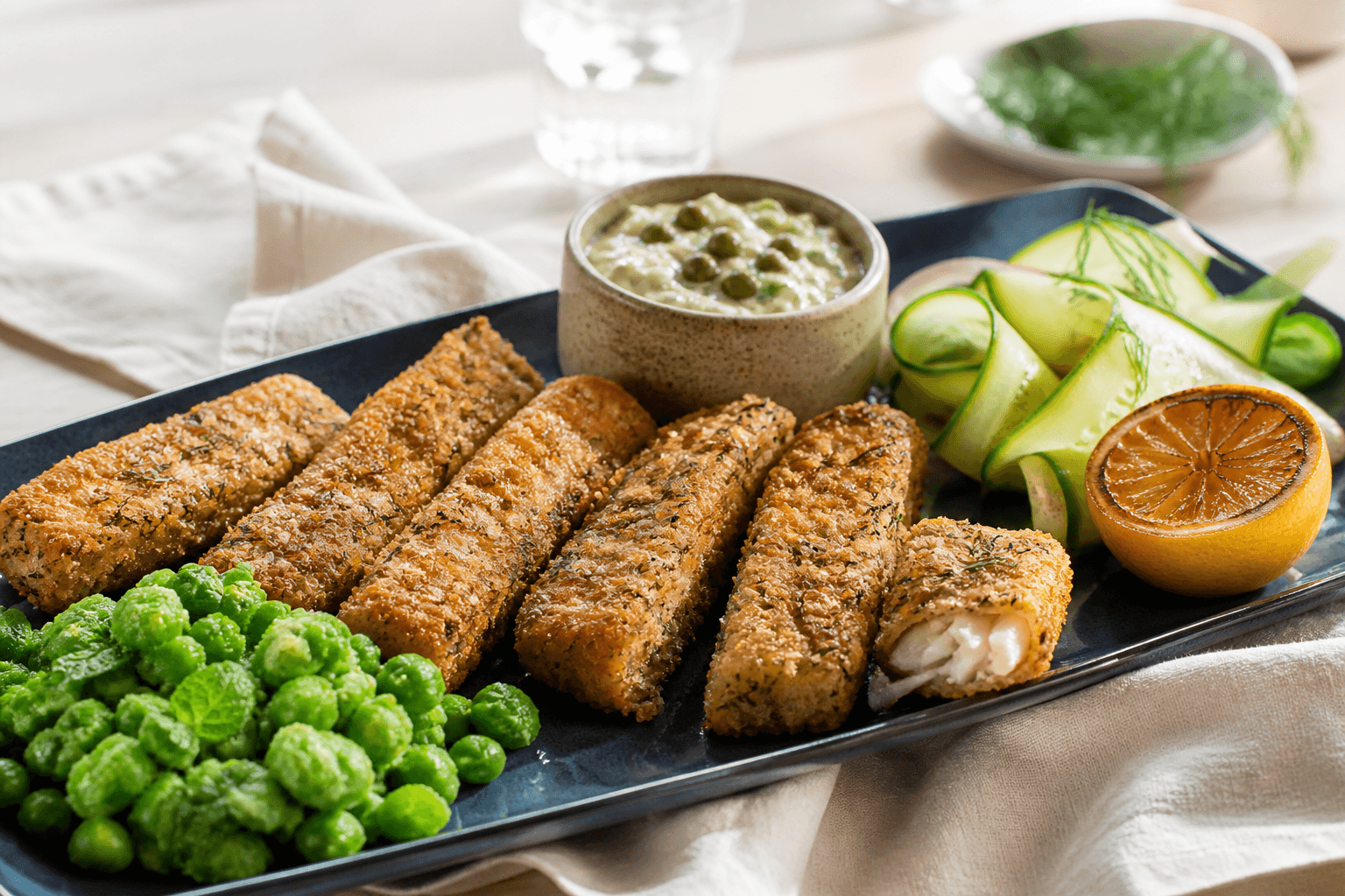 Sprøde rug-panko-fiskefingre med mynteærter, kapers-tartar, syltede agurkebånd og grillet citron på mørkeblåt fad