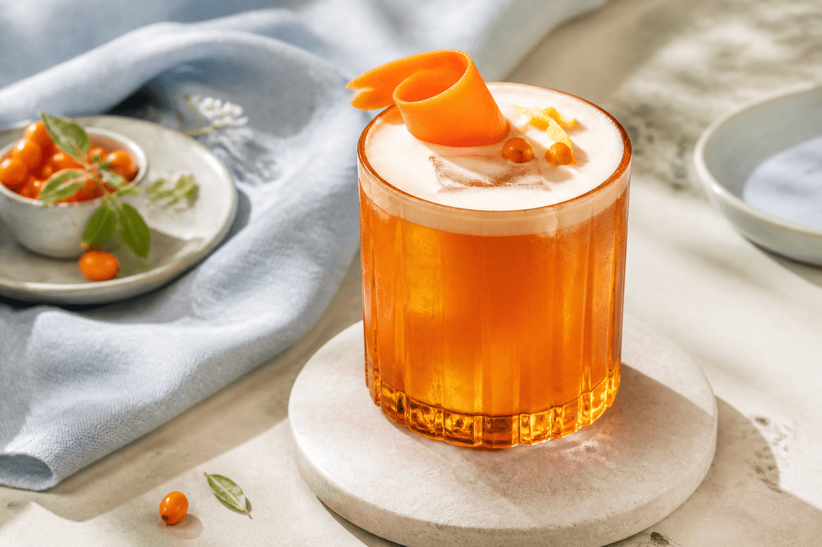 Orange havtorn-gulerod-gin sour med silkeblødt skum, gulerodsbånd, havtorn og citronskal i facetteret ravfarvet glas