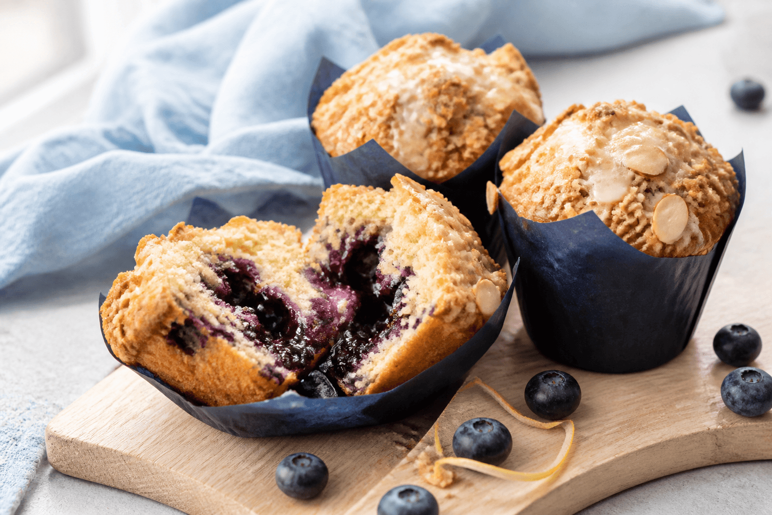 Tre høje citron-blåbærmuffins i indigoblå tulipanforme med marcipan-havrecrumble, mandler, citronglasur og en åben muffin med blåbærkompotkerne