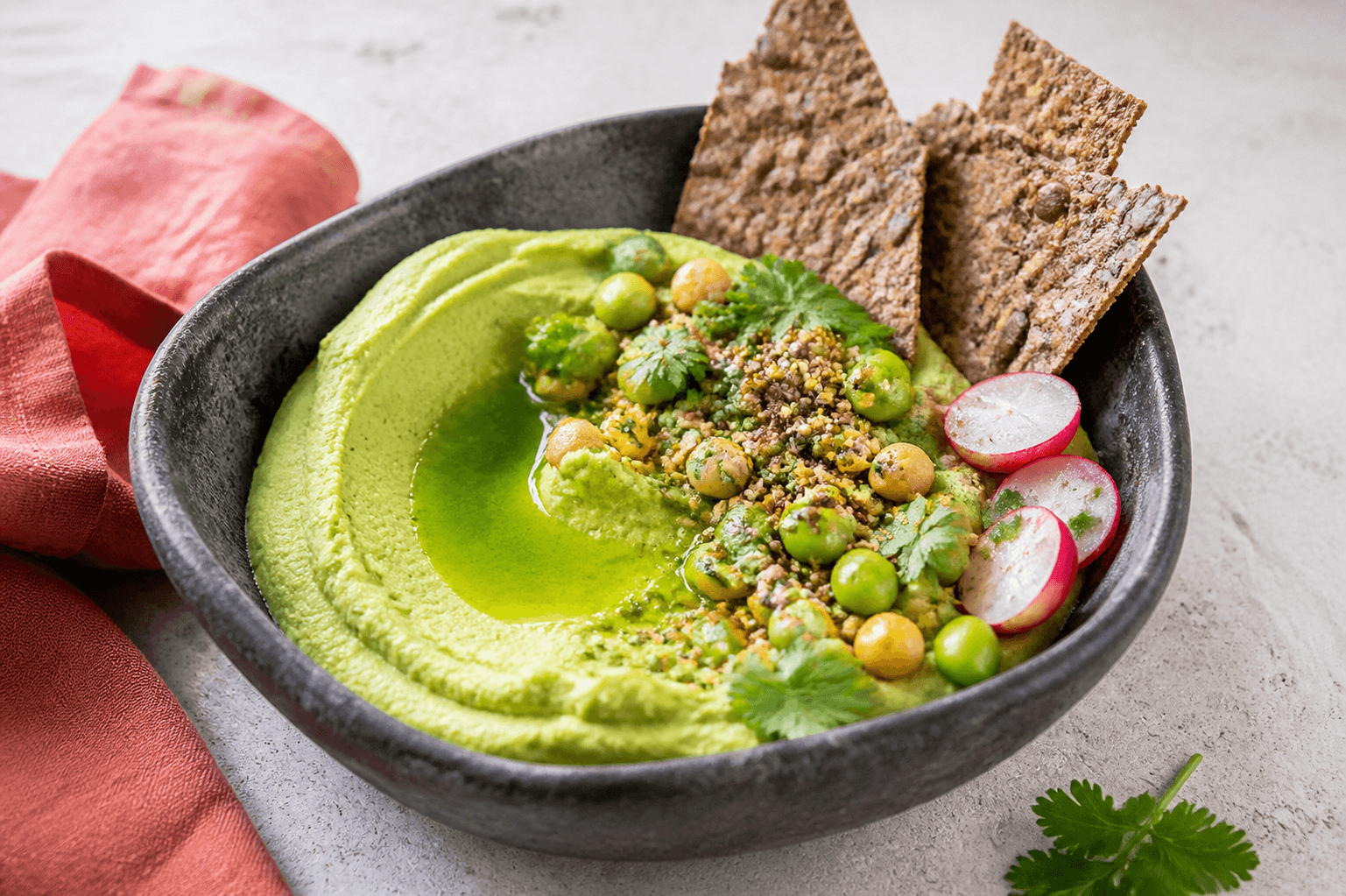 Klargrøn avocado-edamamehummus i mørk stenskål med korianderolie, græskarkerne-dukkah, edamame, ristet majs, radise og sprødt rugfladbrød