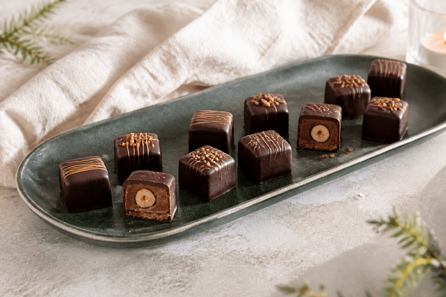 Elleve firkantede hasselnøddetrøfler med mørk chokoladeskal, silkeblød praline, hel ristet hasselnød, rugknas og bronzestriber på mørkegrønt fad