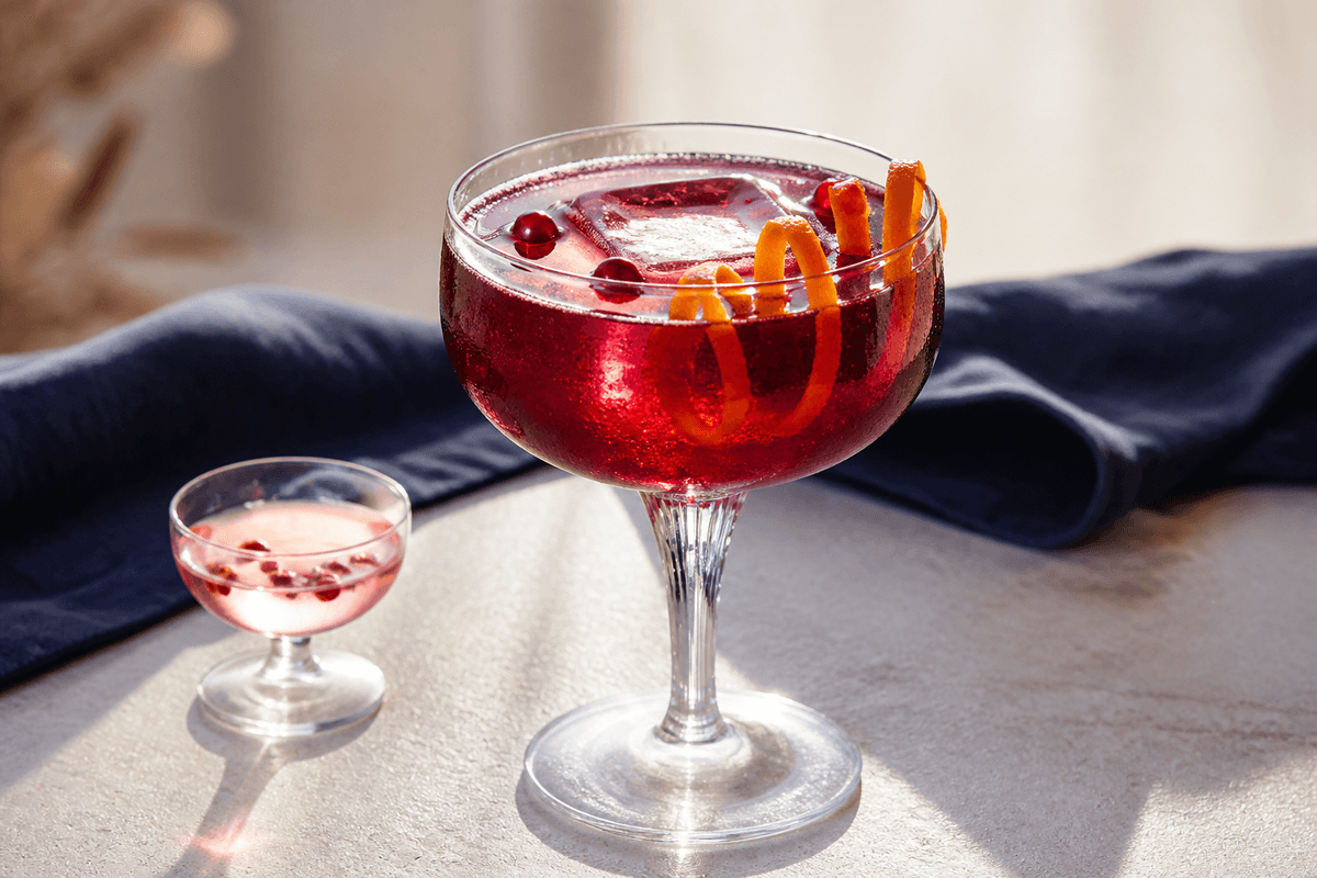 Stilket glas med klar dyb granatæble-blodappelsin-gincocktail, stor klar is, fine bobler, appelsinspiral og granatæblekerner med lille cordialglas