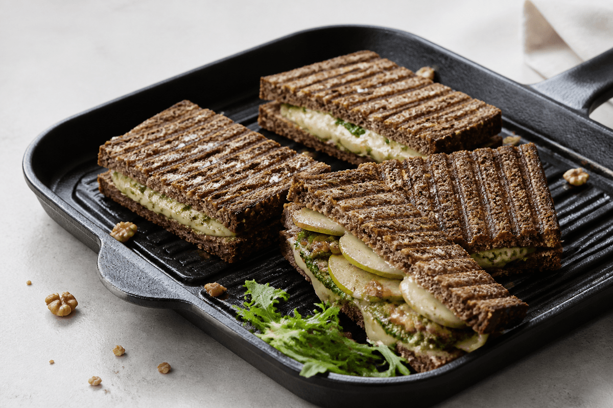 Fire pressede rugbrødstoast med grillstriber i sort grillpande, én åbnet med smeltet brie, tynde pæreskiver, valnødde-persillepesto og karamelliseret løg