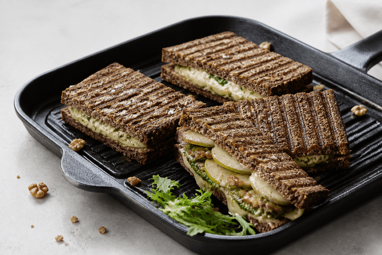 Fire pressede rugbrødstoast med grillstriber i sort grillpande, én åbnet med smeltet brie, tynde pæreskiver, valnødde-persillepesto og karamelliseret løg
