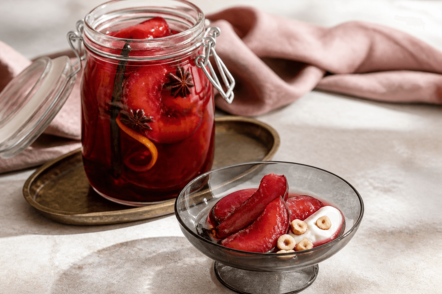 Åbent klart patentglas med rubinrøde råsyltede blommekiler, vanilje, stjerneanis og appelsinskal på messingbakke samt serveringsskål med creme fraiche og hasselnød