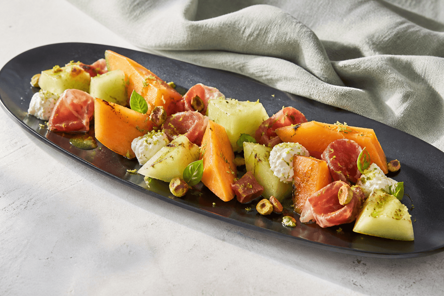 Langt sort fad med cantaloupe og honningmelon, lufttørret skinke i løse bånd, pisket ricotta, pistacie, basilikumolie og lime