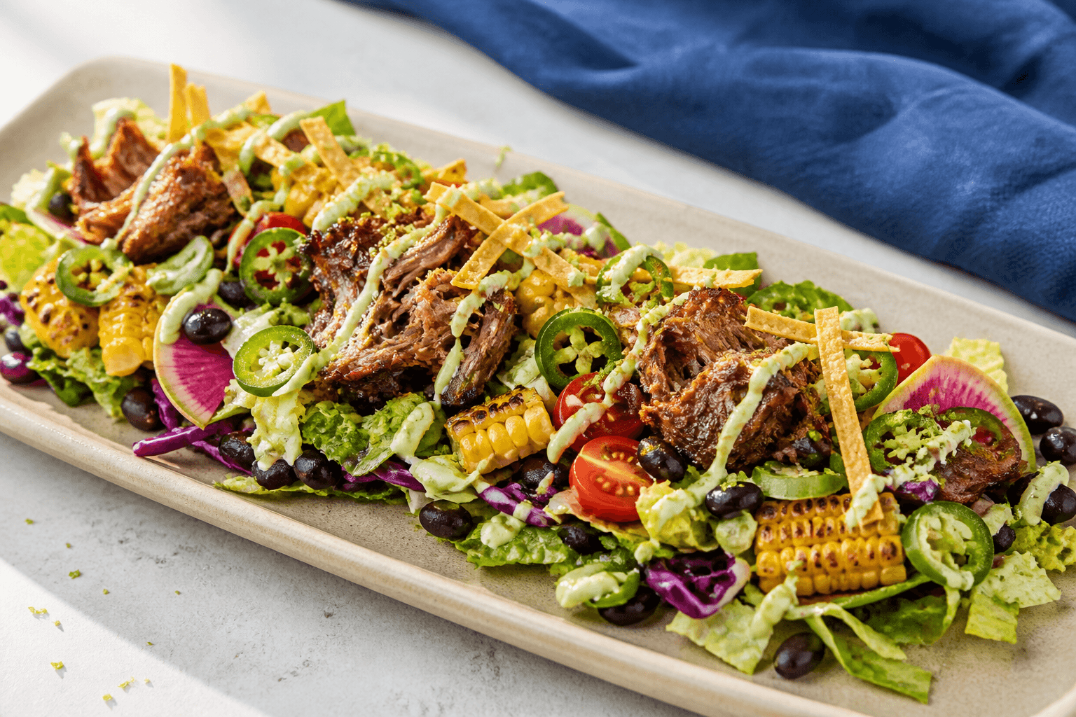 Langt sandfarvet fad med salat, mørkstegt pulled pork, grillede majsstykker, sorte bønner, jalapeño, vandmelonradise, tomat, tortillastrimler og avocado-limecreme
