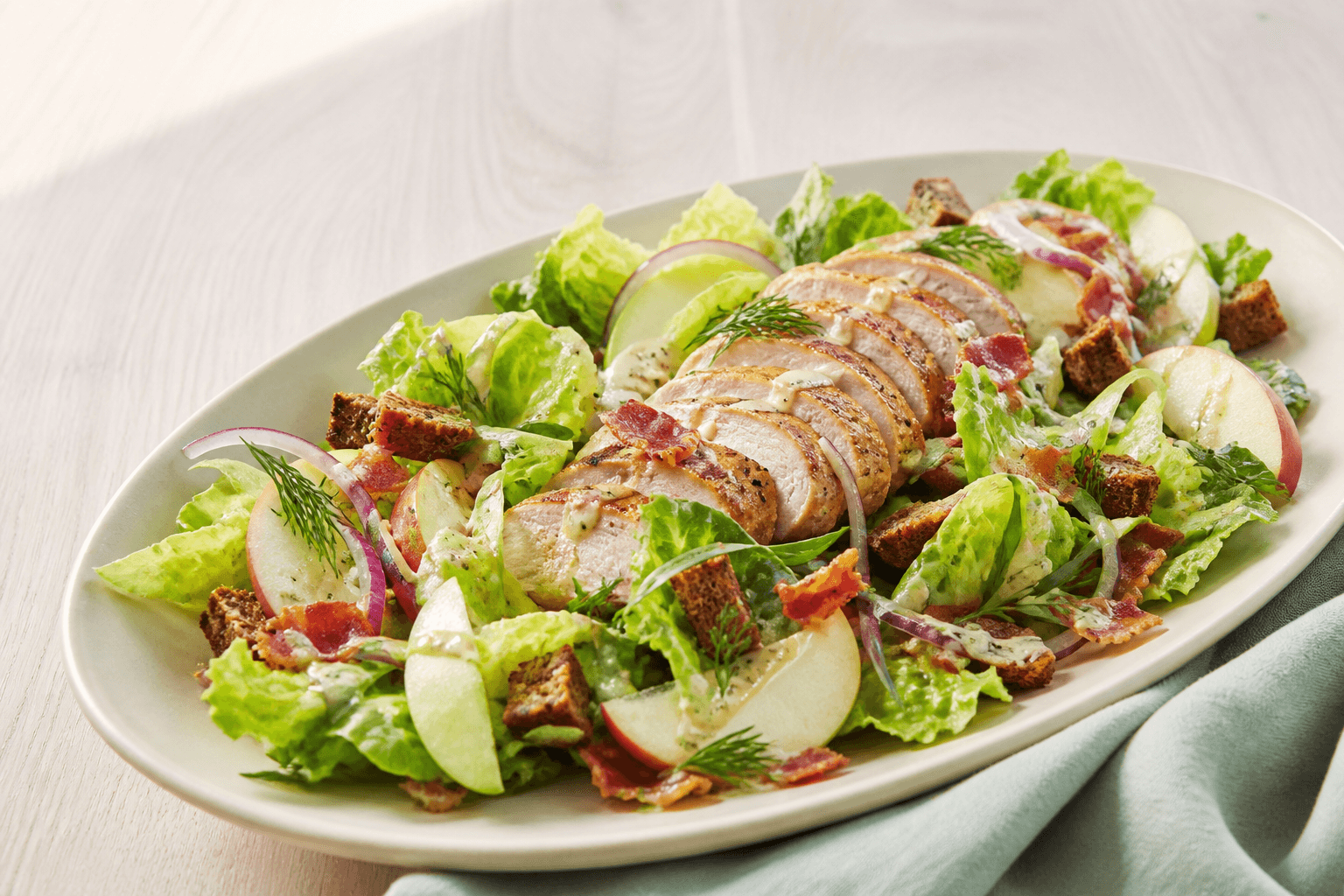 Langt lyst fad med skiver af saftig gylden kylling, little gem, røde og grønne æbler, sprød bacon, rugcroutoner, rødløg, dild og estragon