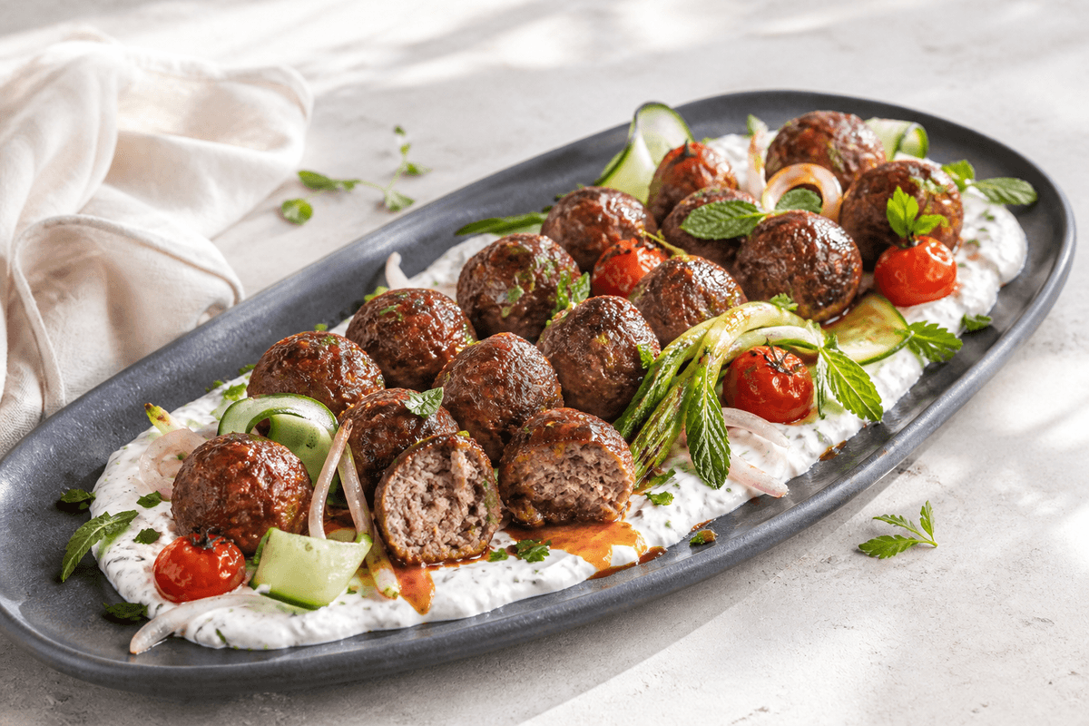 Langt mørkt fad med gyldne lammefrikadeller på citron-mynteyoghurt med aprikos-harissaglace, tomater, forårsløg, agurk og urter