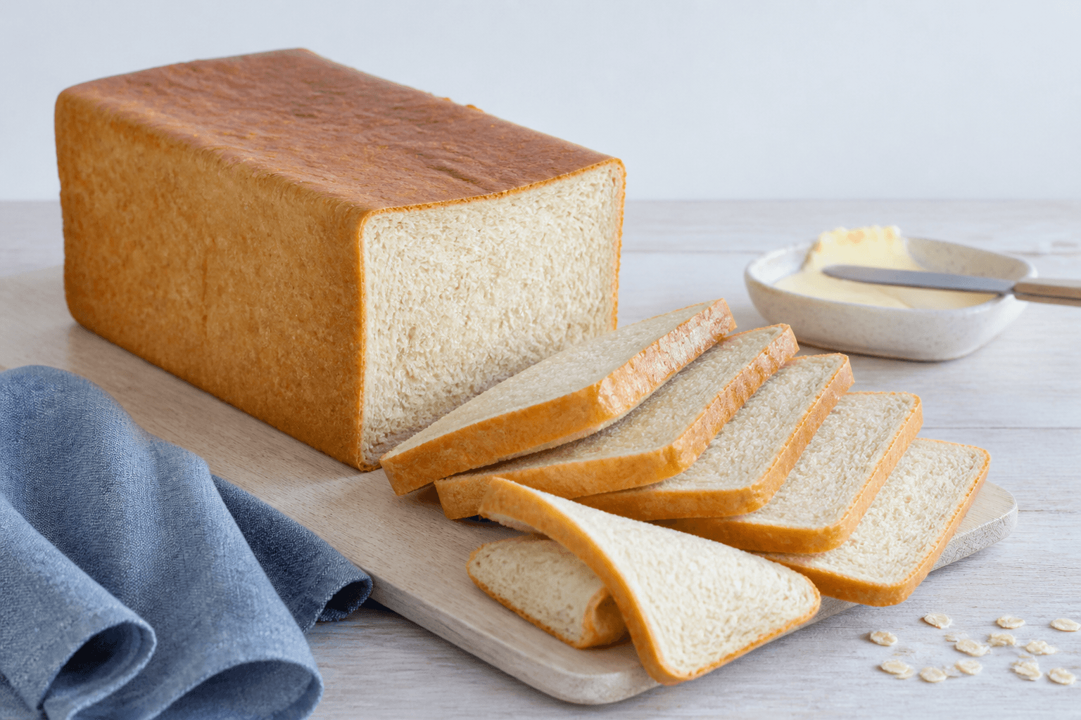 Højt firkantet toastbrød med gylden skorpe, seks ens skiver, fin elastisk krumme, smør og havre på lyst træ