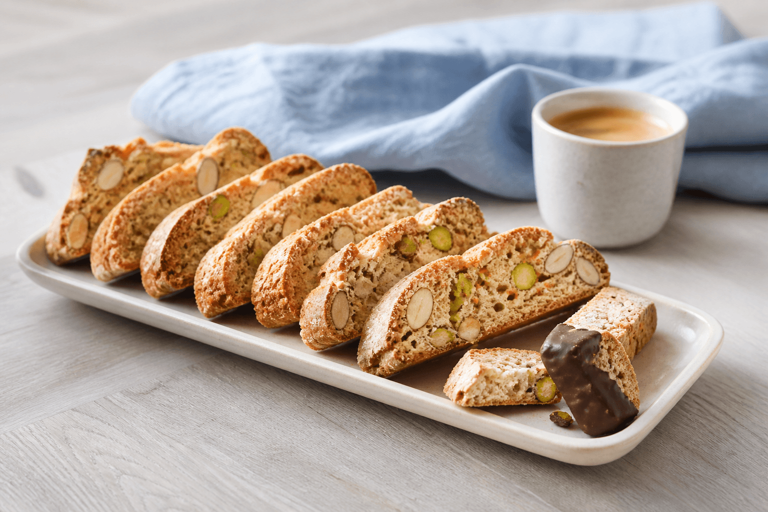 Tynde gyldne cantuccini med hele mandler, pistacie og appelsin på langt cremefad, én brækket og to let dyppet i mørk chokolade