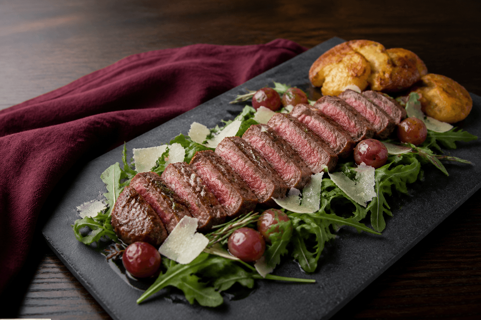 Skiferfad med skiver af rosastegt tagliata med mørk skorpe, rucola, parmesan, ristede druer og sprøde knuste kartofler