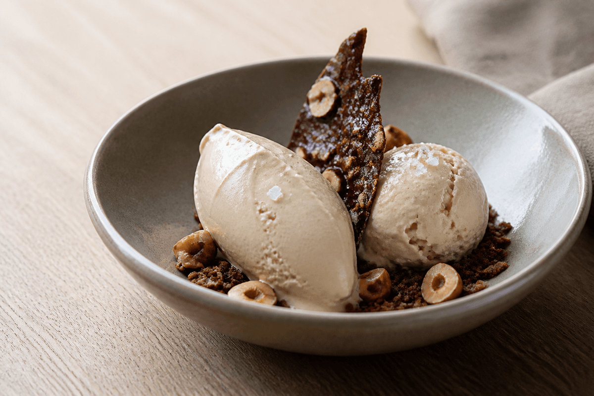 Cremet beige hasselnøddeis som quenelle og kugle i grå skål med chokoladebrittle, ristede hasselnødder og mørk crumble