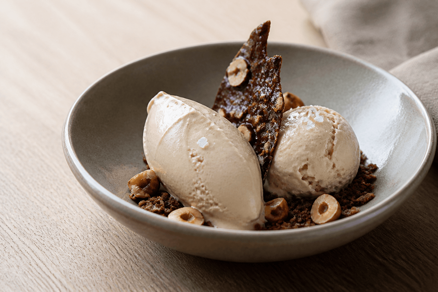 Cremet beige hasselnøddeis som quenelle og kugle i grå skål med chokoladebrittle, ristede hasselnødder og mørk crumble