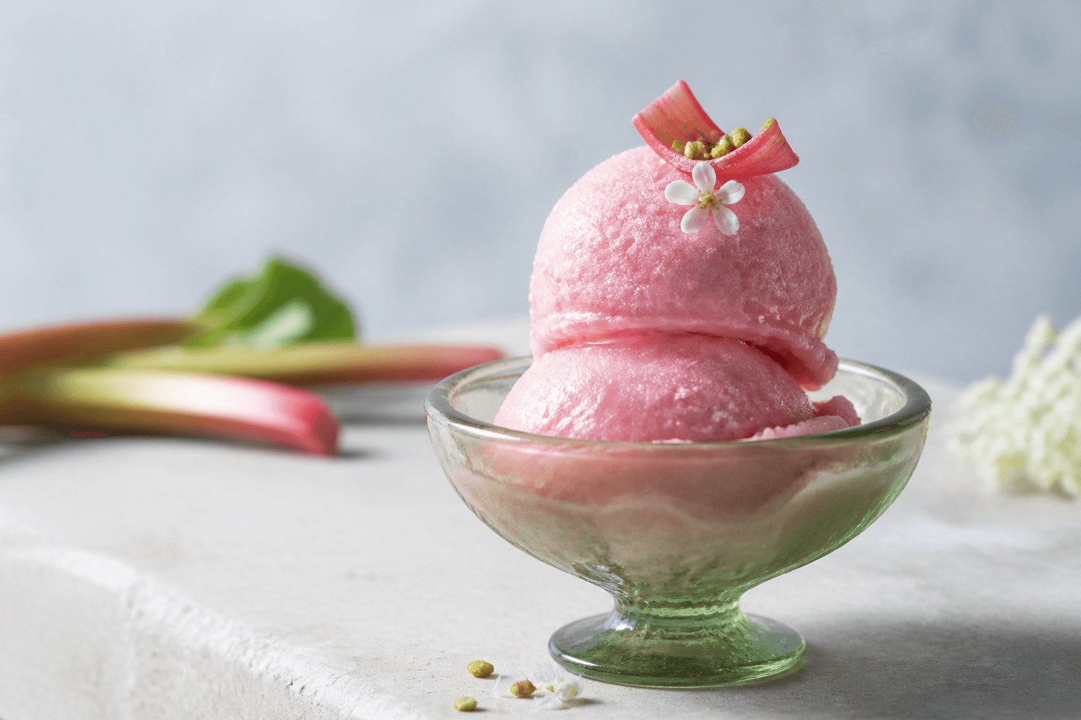 To rosa kugler rabarber-hyldeblomstsorbet i grøn glascoupe med pocheret rabarberbånd, pistacie og en lille blomst