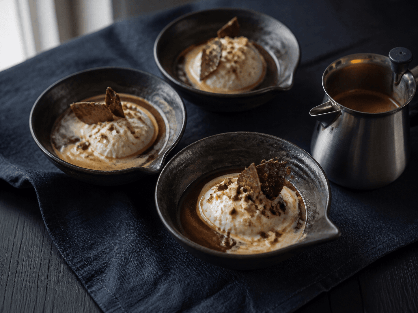 Tre mørke stentøjsskåle med vanilje-skyris, frisk espresso, kakao-rugbrittle og ristede hasselnødder ved en lille stålkande