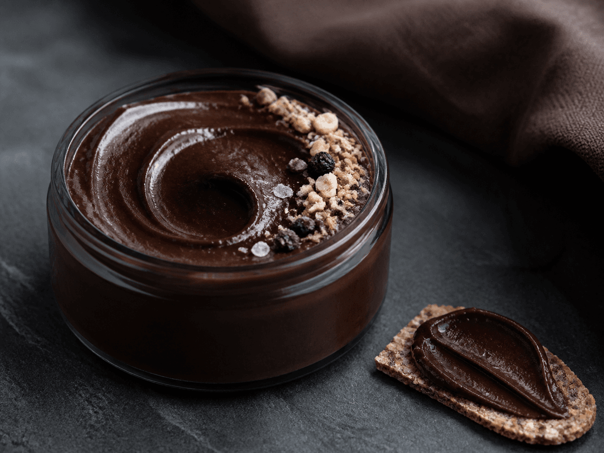 Lav røggrå krukke med blank mørk chokoladecreme, rugmalt, kaffe, ristede hasselnødder, kakaonibs, solbær og flagesalt samt et rugknækbrød