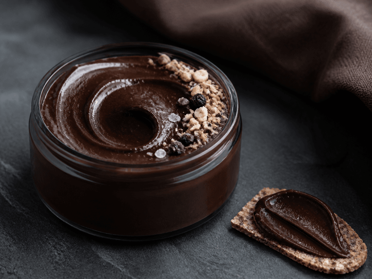 Lav røggrå krukke med blank mørk chokoladecreme, rugmalt, kaffe, ristede hasselnødder, kakaonibs, solbær og flagesalt samt et rugknækbrød