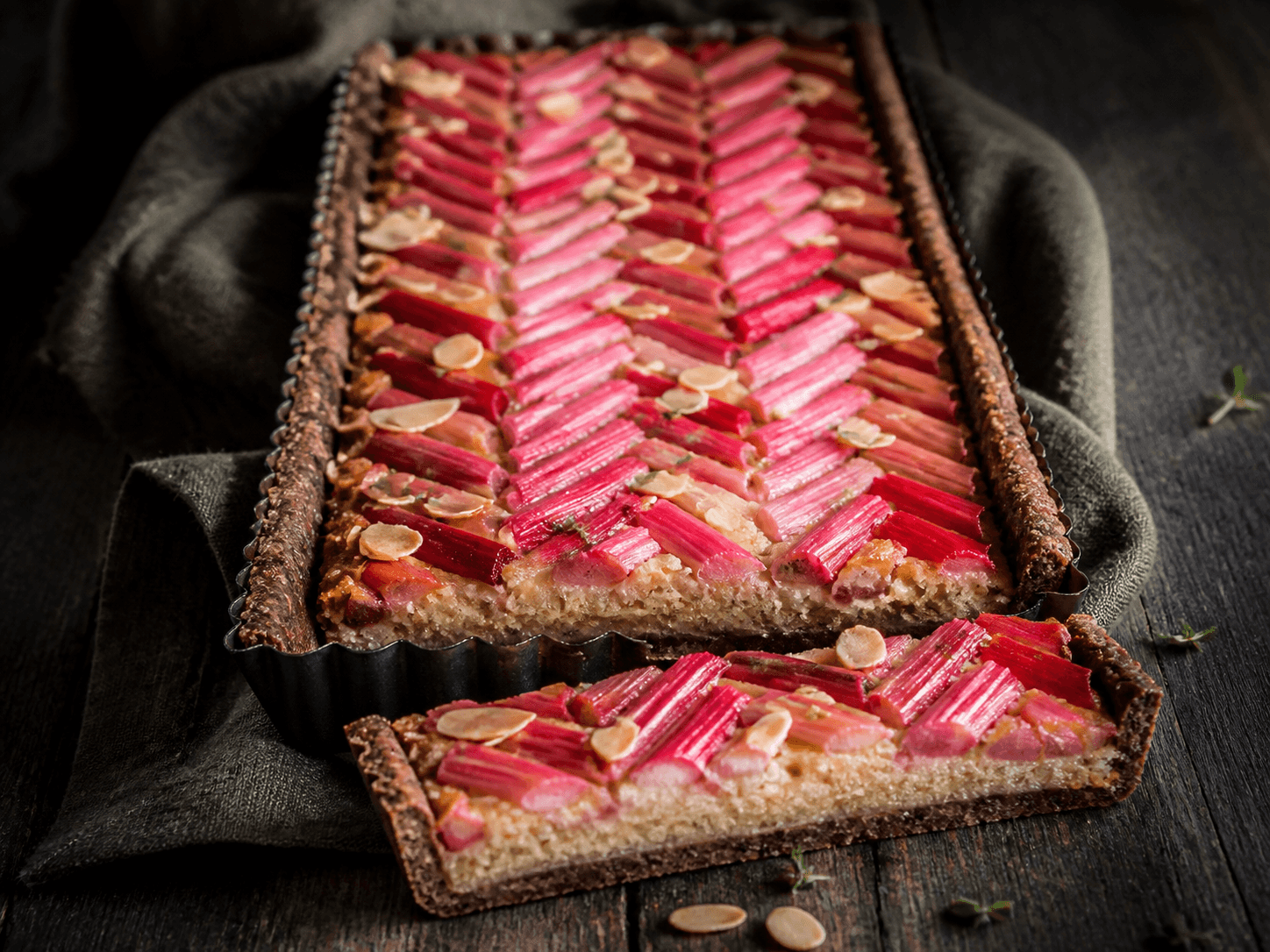 Rektangulær rabarbertærte med mørk rugmørdej, gylden mandelfrangipane, røde rabarber i sildebensmønster, mandelflager og citrontimian