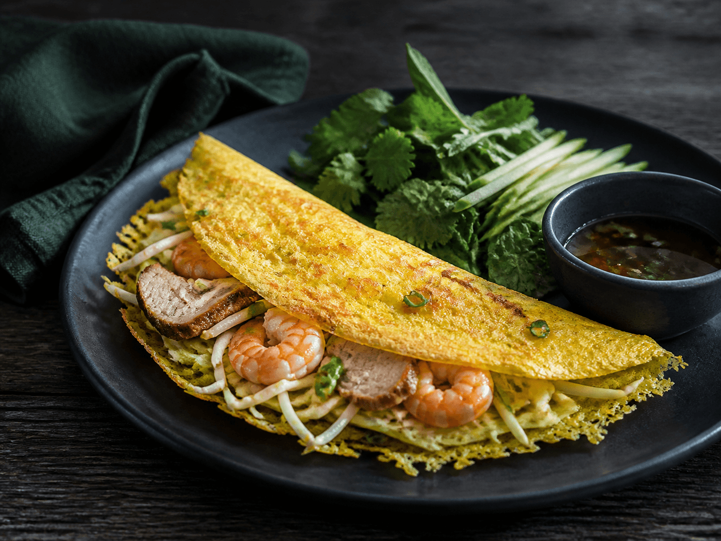 Ultratynd sprød gul banh xeo på sort tallerken med rejer, brunet svinekød, spirer, kålrabi, grønt æble, koriander, mynte og klar lime-fiskesaucedip