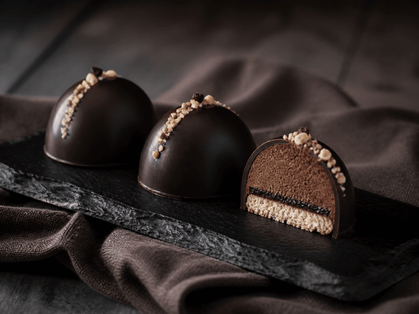 Tre lave mørke Sarah Bernhardt-kupler på sort skifer med hasselnød, espresso-rugmalttrøffel, tyndt solbærlag og blank tempereret chokoladeskal