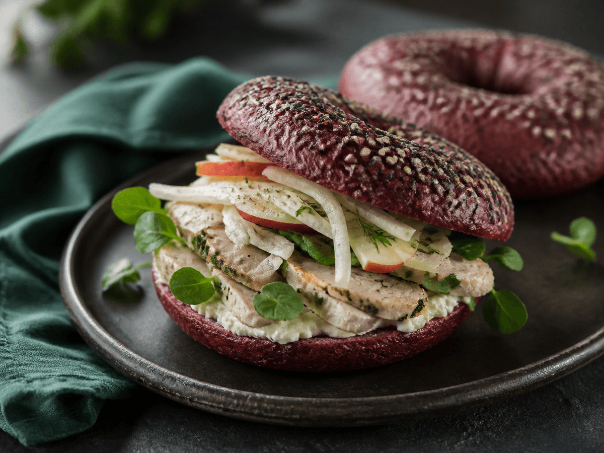 Mørk rødbede-rugbagel med gennemstegt urtekylling, peberrodsflødeost, æble-fennikelslaw og brøndkarse på sort tallerken