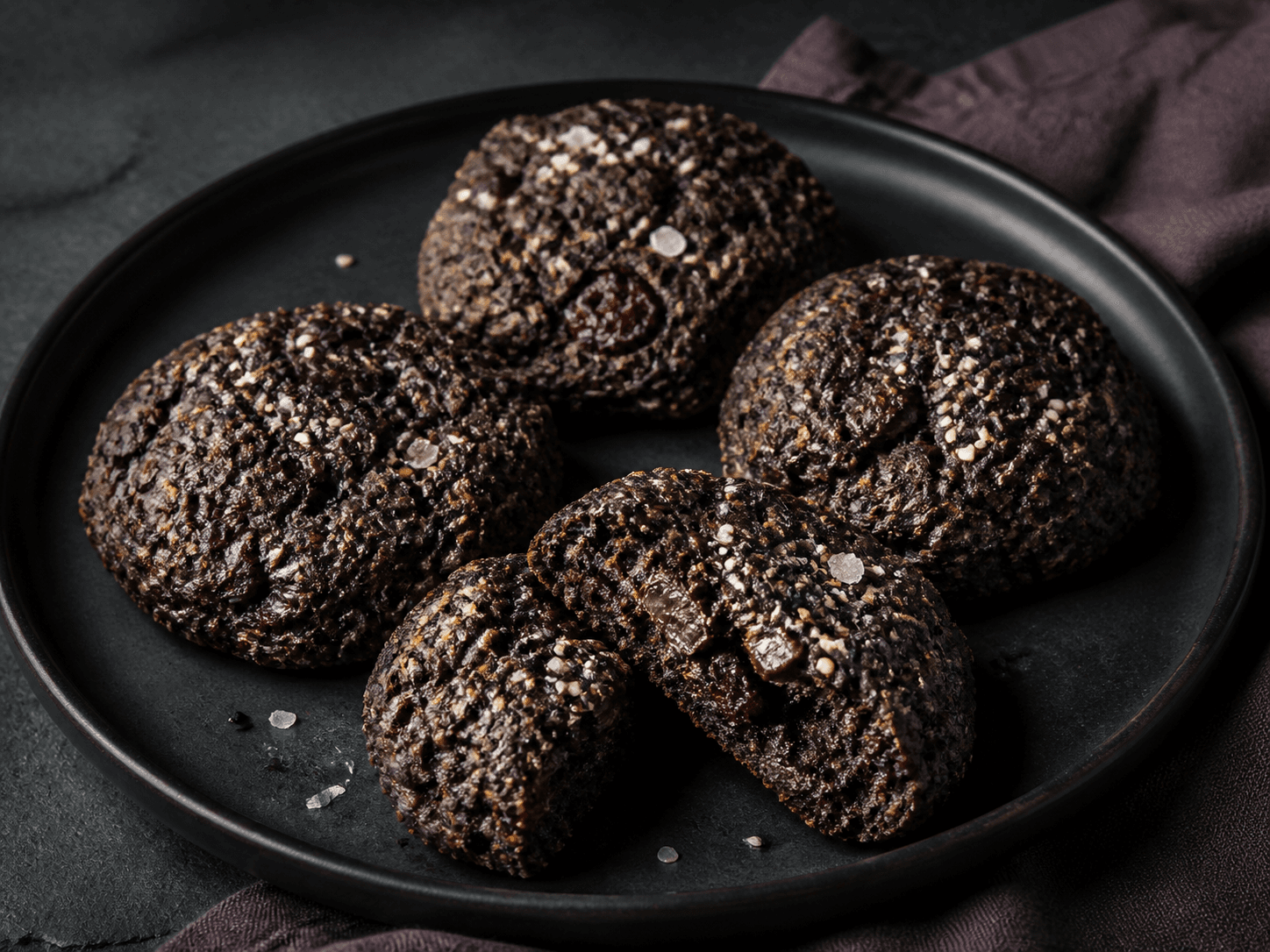 Fire mørke sorte bønnecookies med tahin, mørk chokolade, surkirsebær, sesam og flagesalt på mat sort tallerken