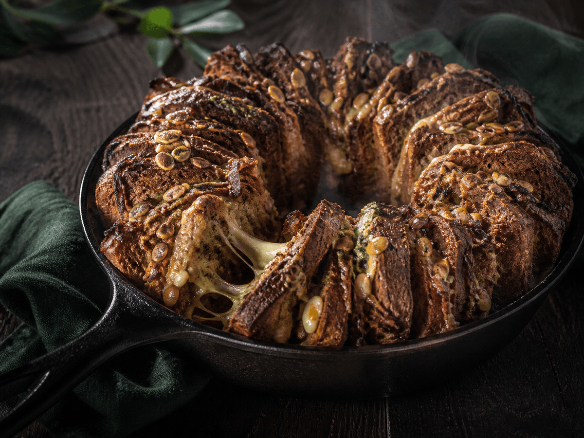 Rundt mørkt rug-spelt pull-apart-brød i støbejern med smeltet lagret ost, karamelliserede løg, ramsløgssmør og græskarkerner