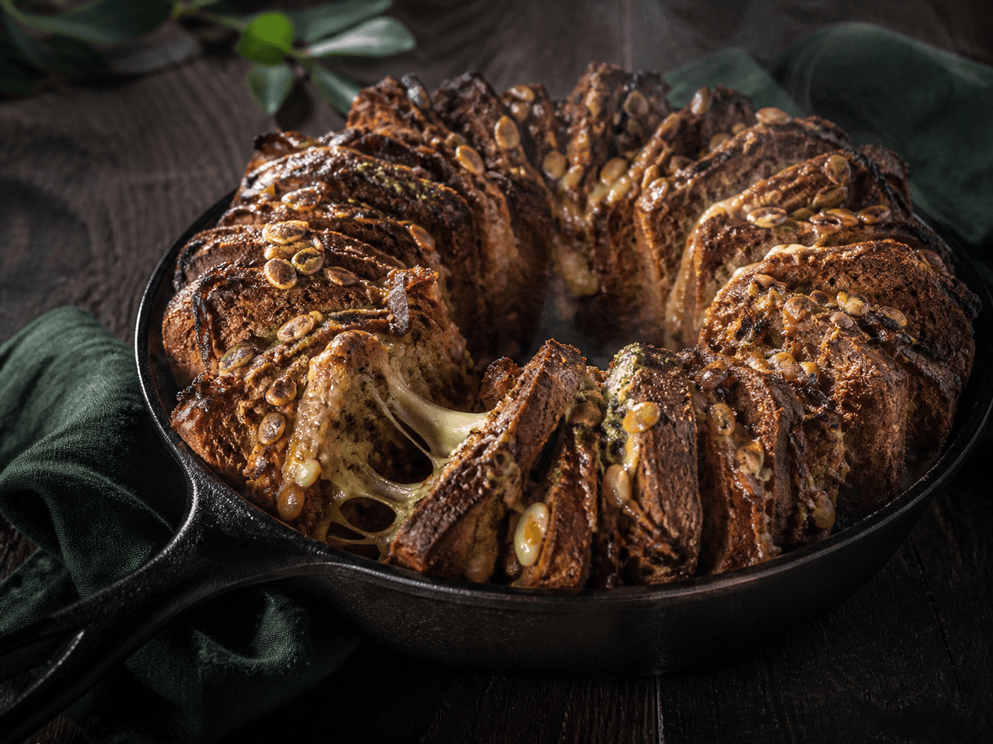 Rundt mørkt rug-spelt pull-apart-brød i støbejern med smeltet lagret ost, karamelliserede løg, ramsløgssmør og græskarkerner