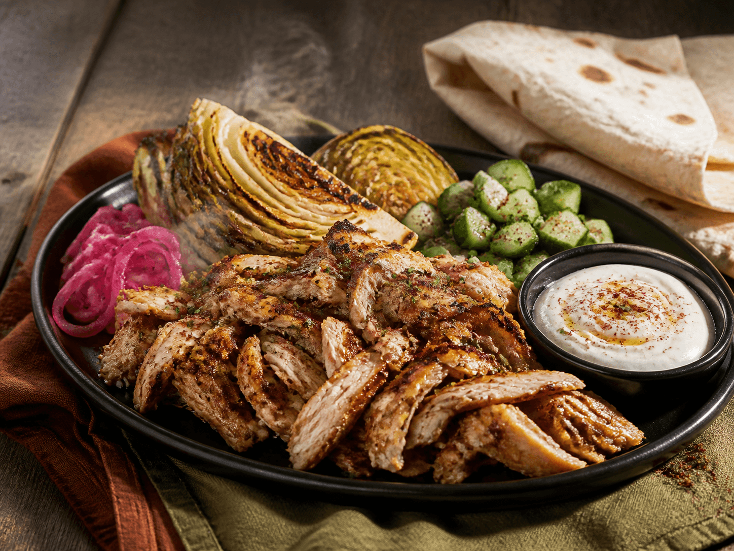 Mørkt fad med sprødkantet gennemstegt kyllingeshawarma, bagt spidskål, syltet rødløg, agurk, tahin-skyr og bygfladbrød