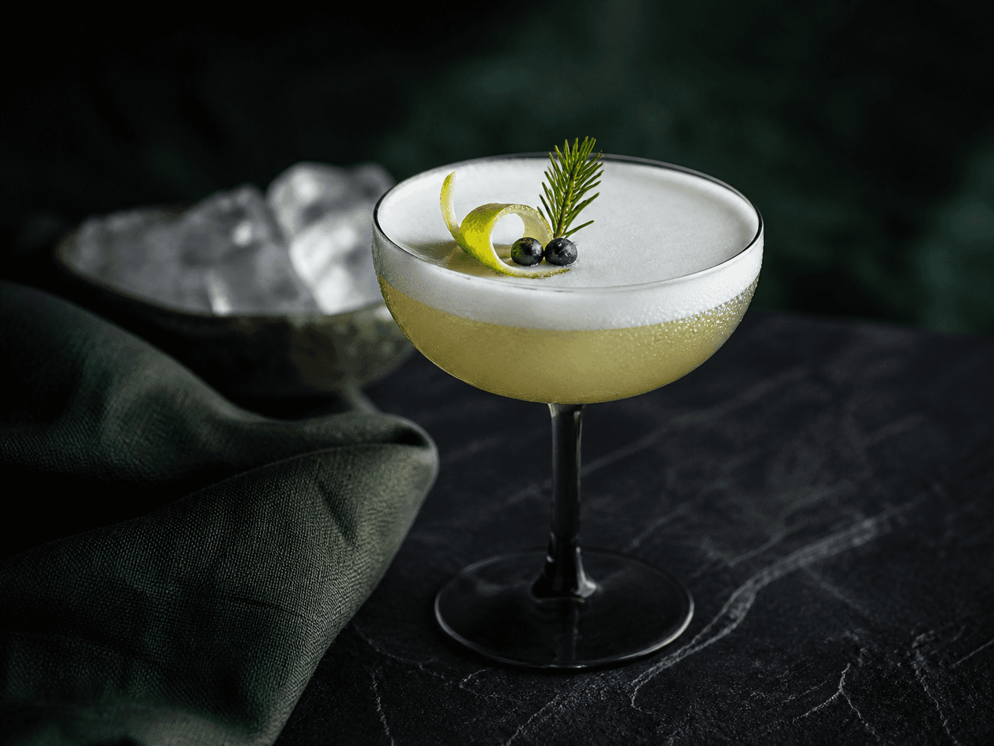Røggrå coupe med lys bergamotte-gin-sour, fint skum, bergamotteskal, enebær og lille granskud på sort sten