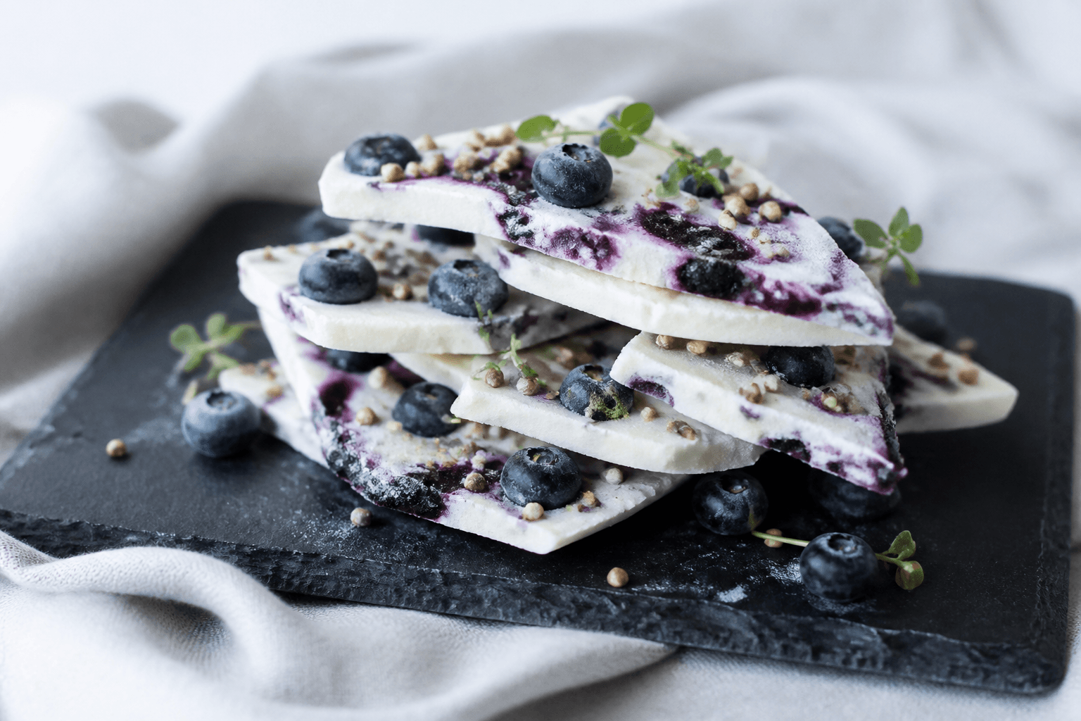 Stak af frozen-yoghurt-bark med blåbærkompot, friske blåbær, honningristet boghvede og citrontimian på mørk skifer