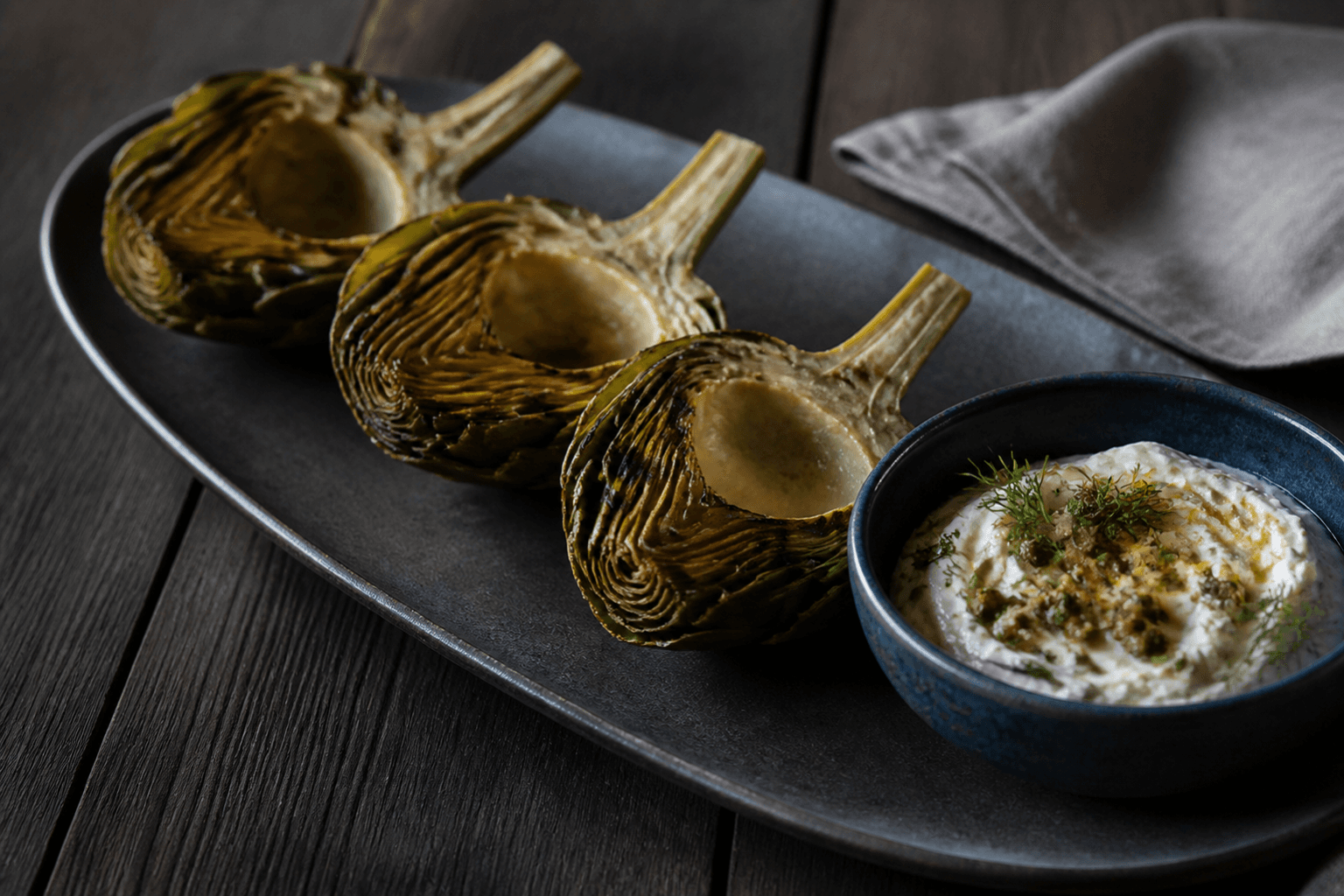 Tre halve mørkt grillede artiskokker med fjernet skæg og en blå skål pisket brunet smør-skyr med dild og sprøde kapers