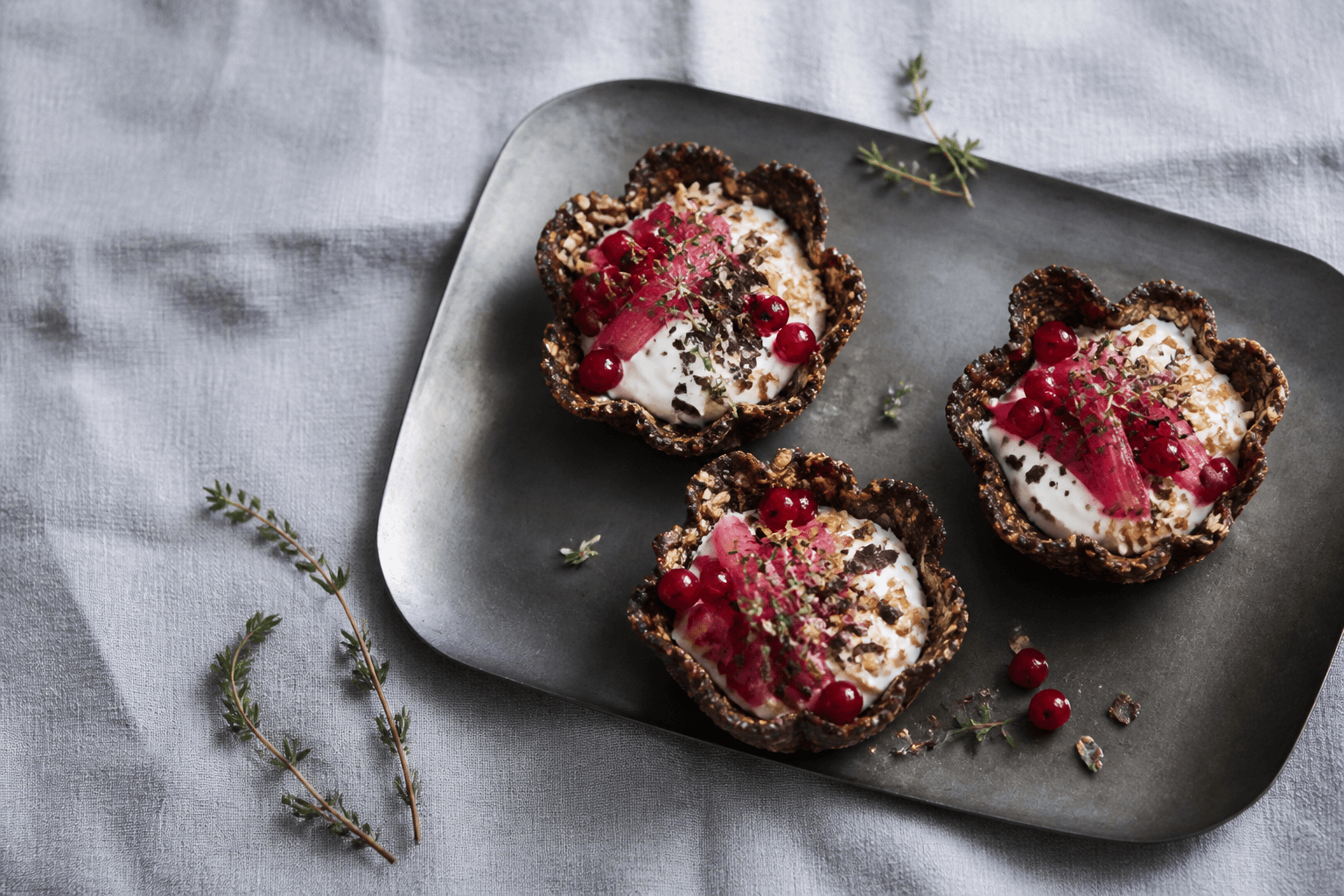 Tre mørke riflede daddel-havreskåle med skyr, ovnbagt rabarber, ribs, havreknas og mørk chokolade på sort metalbakke
