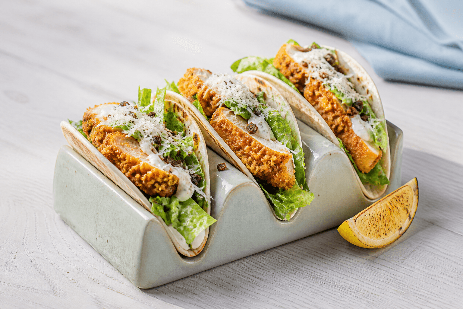 Tre sprøde Cæsar-tacos med gennemstegt paneret kylling, romaine, citrondressing, lagret ost og rugknas i lys salviegrøn holder