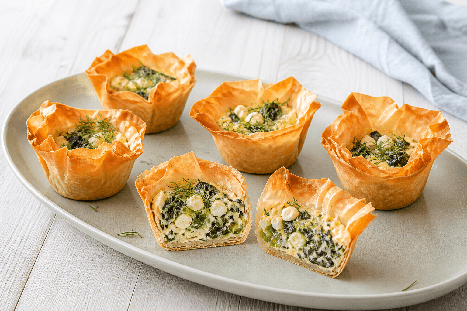 Seks gyldne filodejsmuffins med spinat, porre, feta og dild på langt salviegrønt fad med én skåret over