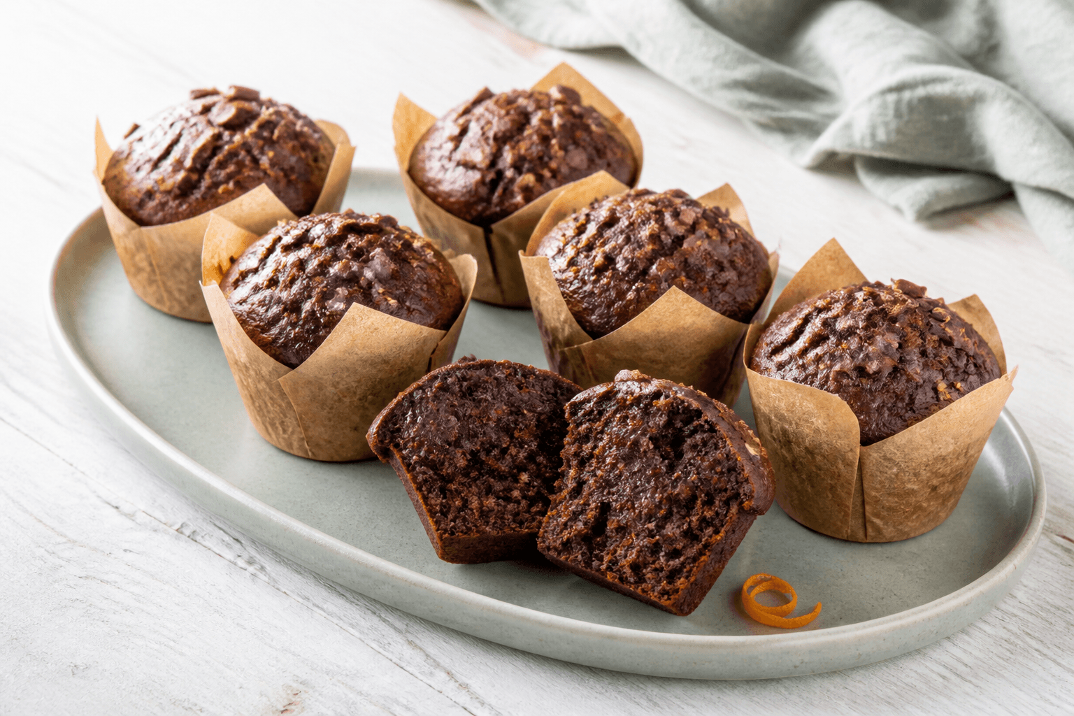 Seks mørke chokolademuffins i brune forme med chokoladestykker, kakaonibs og én åben fugtig krumme på lysegrønt fad