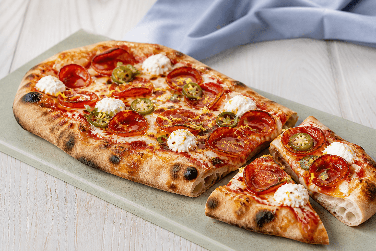 Aflang hot-honey-pizza med luftig bund, sprød pepperoni, jalapeño, ricottaprikker, oregano og tynde honningtråde på salviegrøn sten
