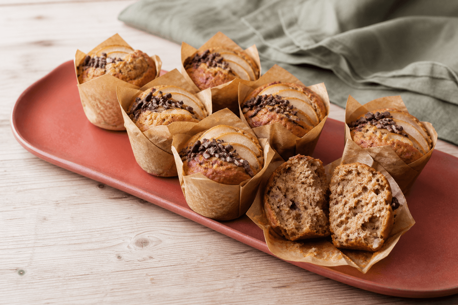 Otte høje veganske dadelmuffins i tulipanpapir med pærevifter, kakaonibs og én åben sat havrekrumme på koralfad