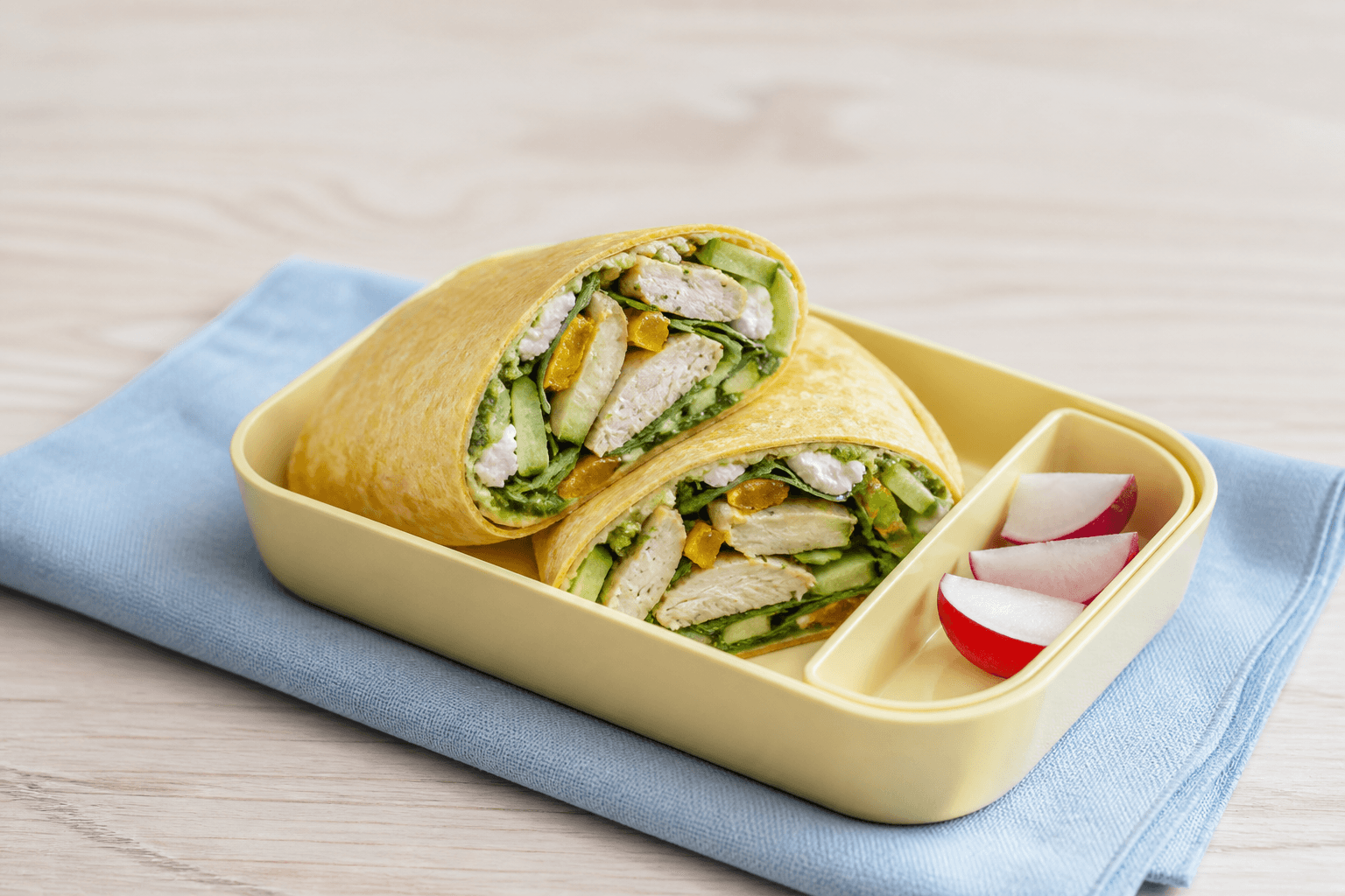To skråt skårne gyldne hytteostwraps i gul madkasse med gennemstegt kylling, grøn pesto, gul peber, agurk og spinat