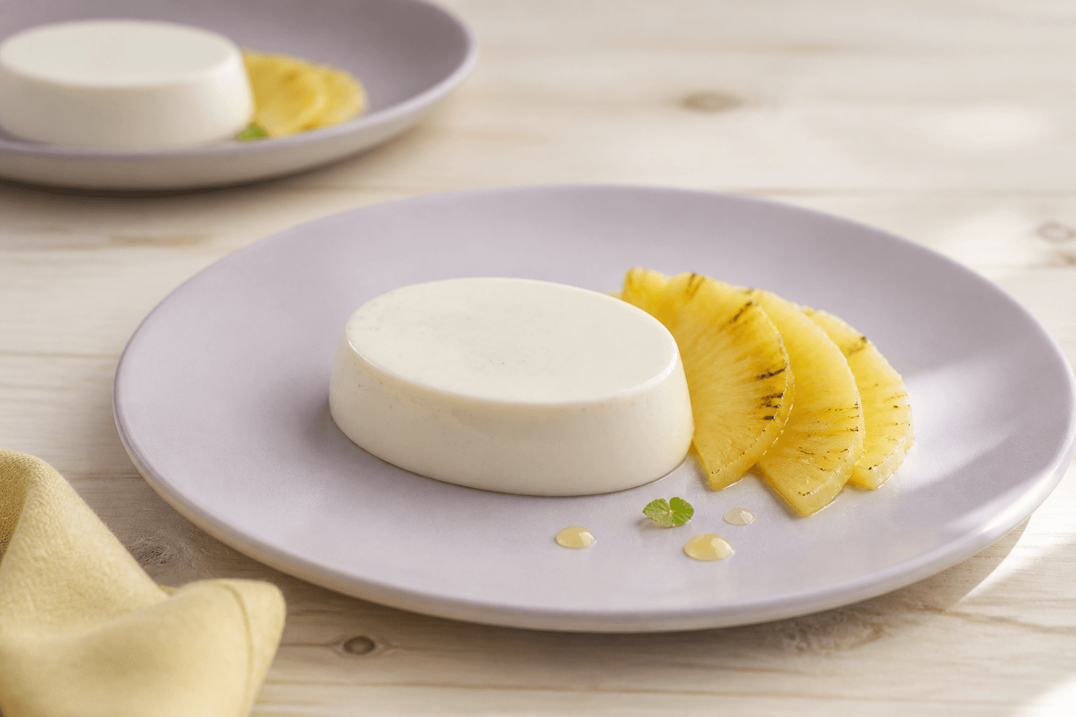 Lav oval vanilje-yoghurt-panna cotta på lavendeltallerken med vifte af tynde grillede ananasskiver og små limedråber