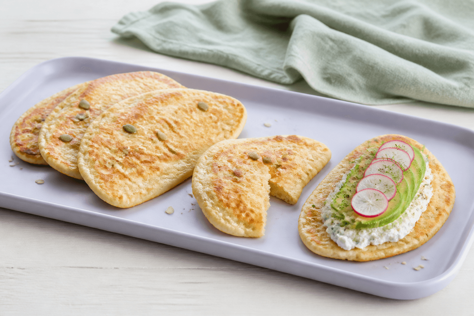 Fire små ovale gyldne hytteost-æg-fladbrød med en smal frølinje på lavendelfad, ét åbent og ét toppet med urtehytteost, avocado, agurk og radise