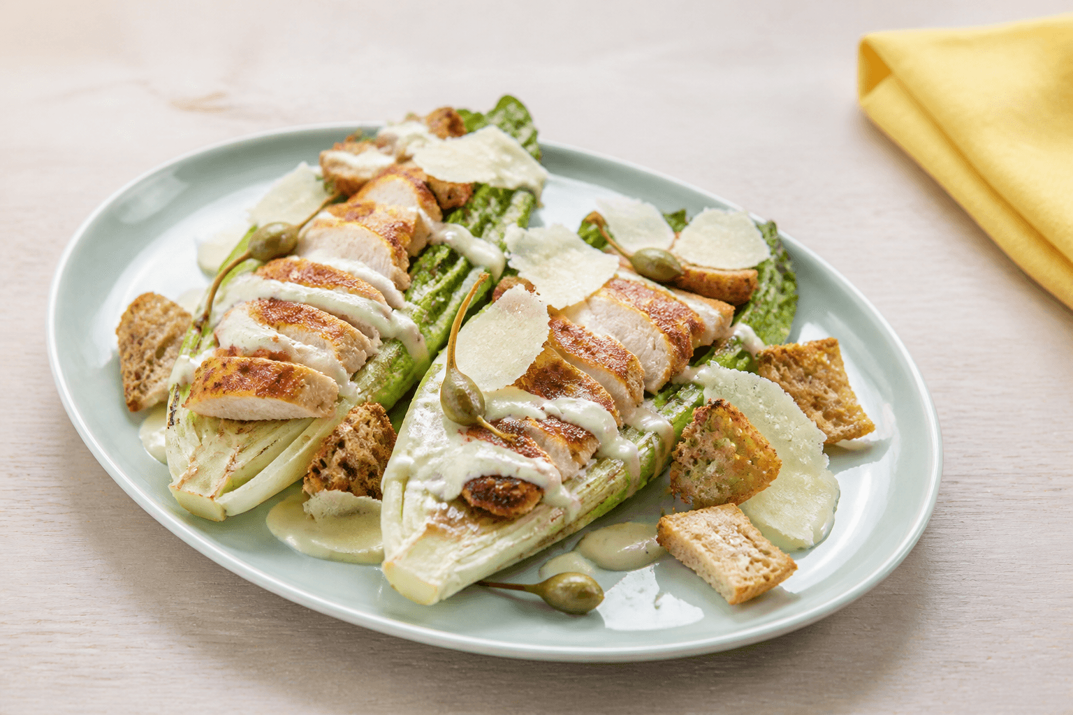 To grillede romainehjerter på lysegrøn tallerken med skiveskåret kylling, rugcroutoner, parmesanspåner, kapersbær og lys cæsardressing