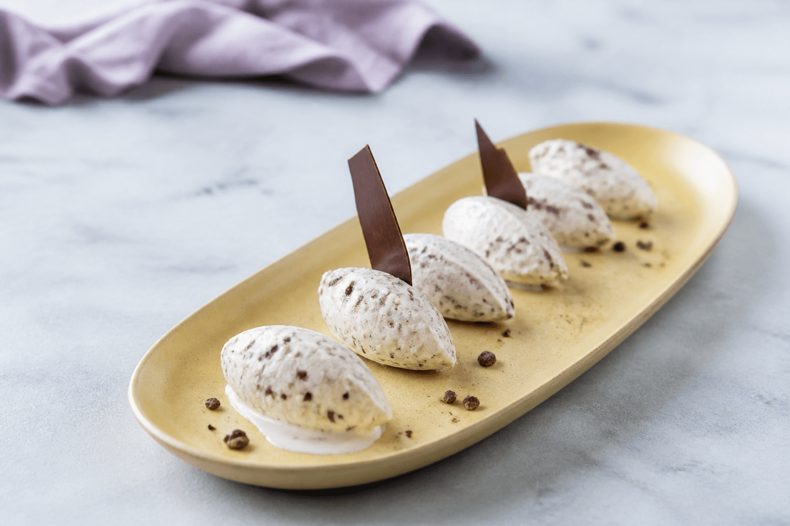 Fem ovale queneller af lys stracciatella-is på smørgult fad med fine mørke chokoladeflager, to chokoladesejl og en lille smeltekant