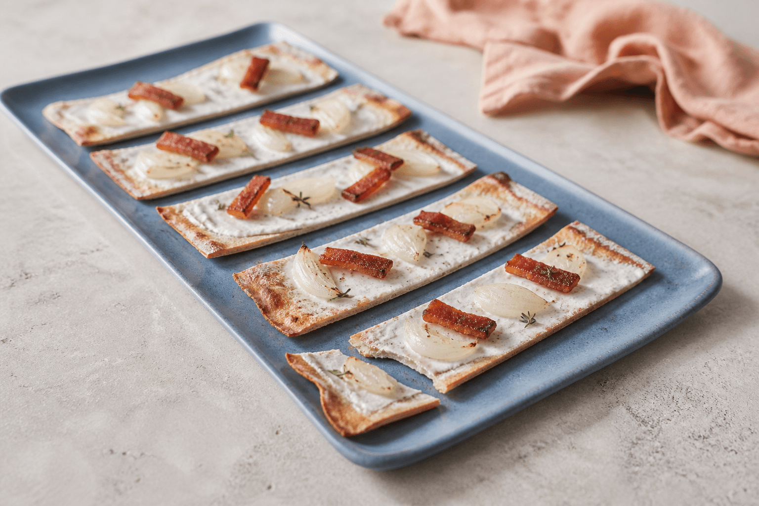 Otte lange sprøde flammkuchenfliser på skiferblåt fad med creme fraiche, ristede løgblade, røget flæsk og timian