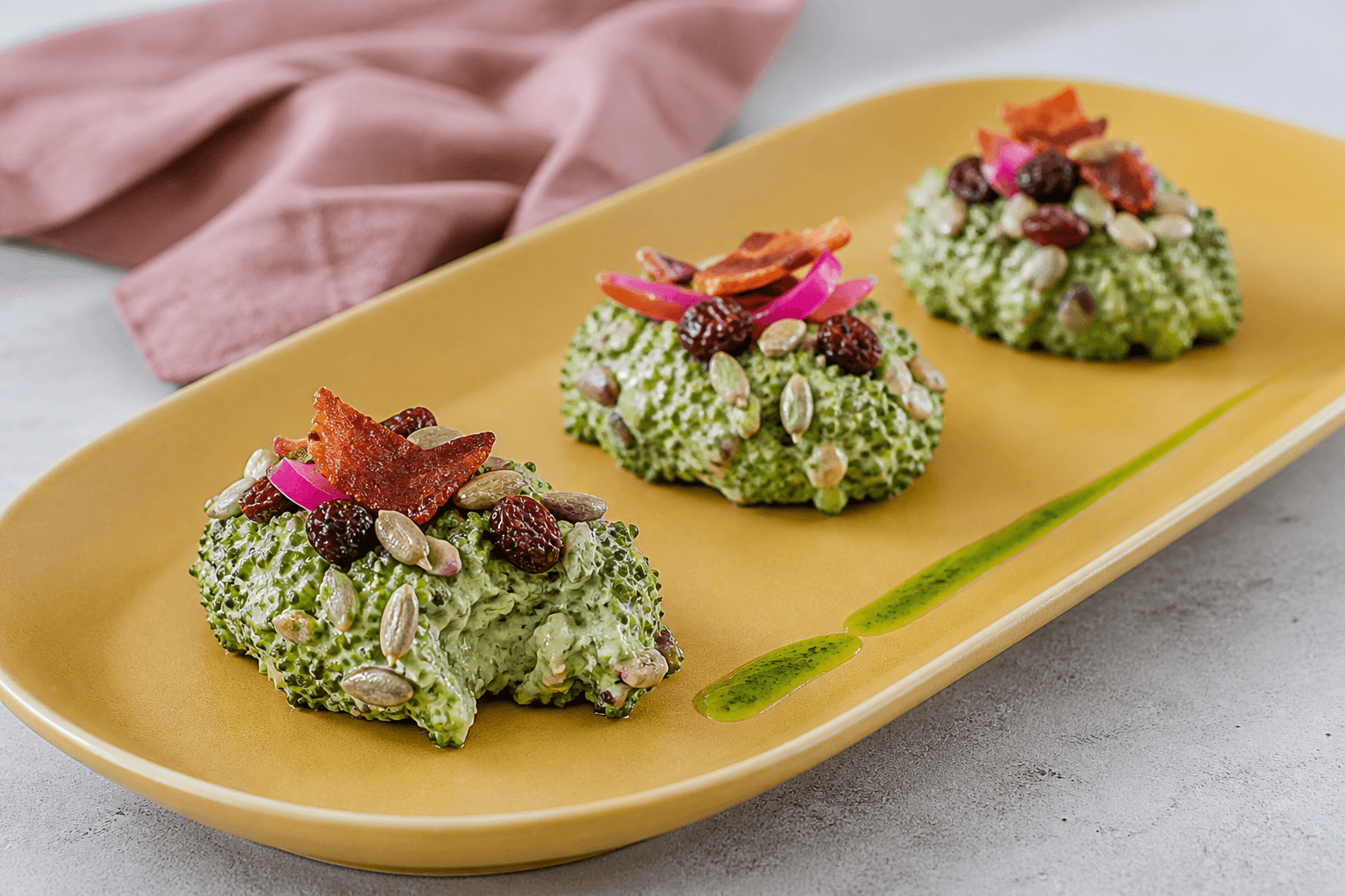 Tre kompakte grønne broccolisalater på sennepsgult fad med sprød bacon, rosiner, syltet rødløg, solsikkekerner og purløgsolie